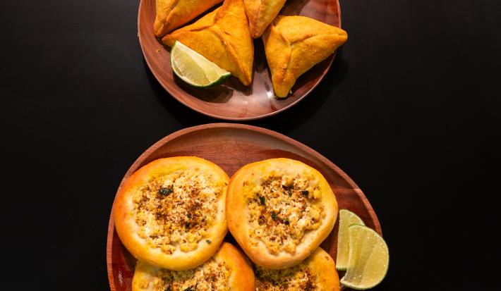 Fotografia em tons de marrom com dois pratos redondos e marrons ao centro. Dentro de um dos pratos existe algumas esfihas fechadas acompanhadas de limão. Dentro de outro prato existe esfihas abertas recheadas de tofu, acompanhadas com limão.