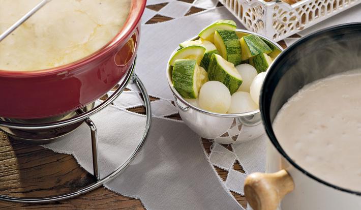 recipientes nas cores vermelho e branco ambos comporta o fondue de queijo, outros recipientes estão com os acompanhamentos para o fondue como pão, presunto entre outros.