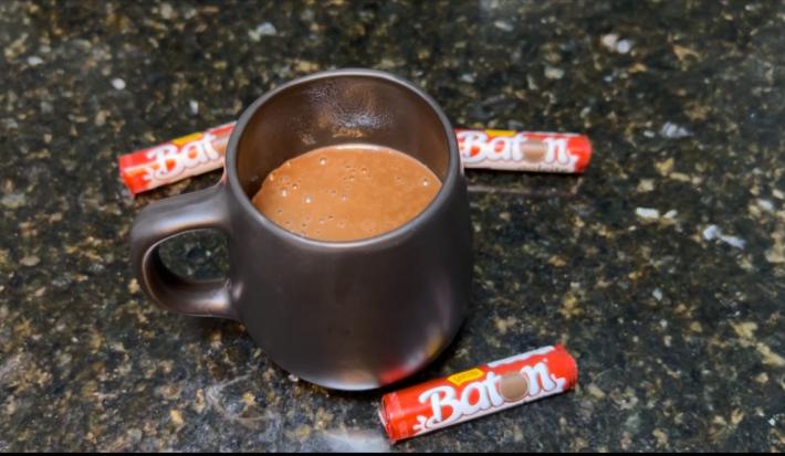 Fotografia em tons de preto com uma caneca preta ao centro. Dentro da caneca existe uma receita de chocolate quente cremosa de cor marrom. Ao lato existe três chocolates em forma de cilindro pequeno da marca BATON