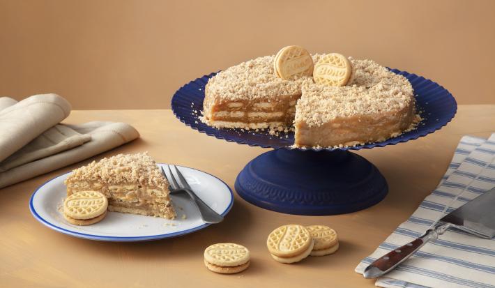 Foto de uma mesa marrom clara, com uma boleira azul escura. Sobre ela está a receita da Torta de Palha Italiana decorada com Biscoitos Bono Doce de Leite. Na mesa há um prato com uma fatia, mais alguns biscoitos Bono e guardanapos de tecido.
