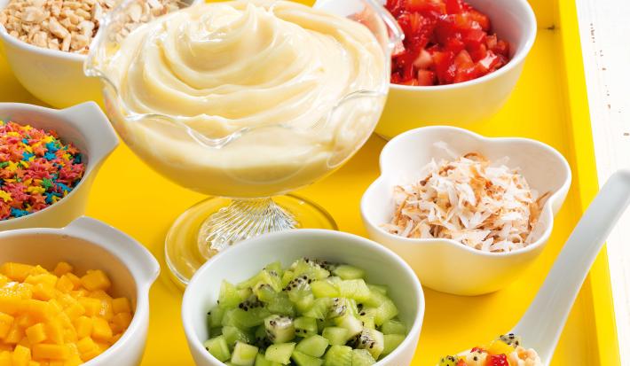 Fotografia em tons de amarelo em uma bancada de madeira clara, uma bandeja amarela com vários potinhos com frutas e doces e ao centro um pote de vidro com o Moça de Colher.