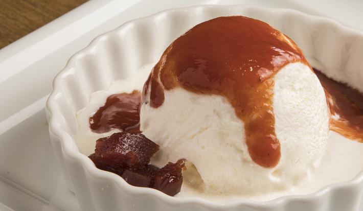 Fotografia de uma bola de sorvete sabor requeijão com uma calda quente de goiaba dentro de uma forma pequena e redonda na cor branca. O recipiente está apoiado em um prato banco quadrado, que está apoiado em uma mesa de madeira.