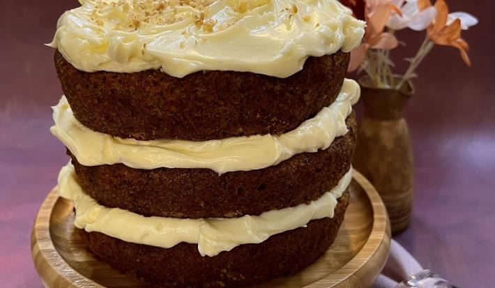 Foto em tons de marrom da receita de carrot cake servida sobre uma tábua redonda de madeira. Ao lado esquerdo há um prato de porcelana com um garfo prateado, ao lado direito há um lenço cor de rosa