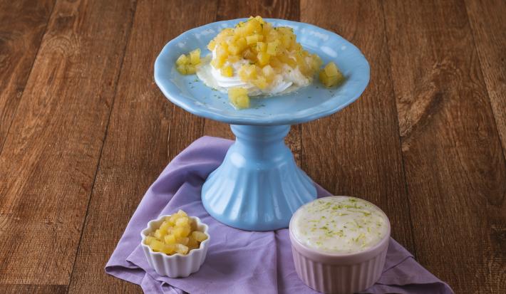 Foto da receita de pavlova de limão com abacaxi, servida em um prato azul, com creme ao lado em um pote rosa e outro pote branco com pedaços de abacaxi, tudo em uma bancada de madeira com um pano em tom violeta