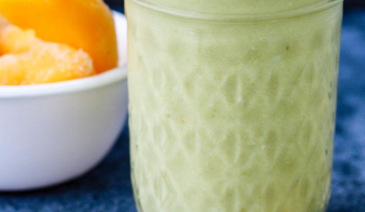 Fotografia em tons de verde em uma mesa de madeira com uma toalha azul de veludo. Ao centro, um copo grande de vidro com o smoothie de matchá e pêssego. Ao lado, uma colher dourada e um potinho com pedaços de pêssego.
