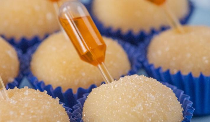 Fotografia de sete forminhas azuis juntas com brigadeiro de pudim, em cada unidade tem uma mini pipeta espetada com calda de caramelo. Os brigadeiros estão apoiados em um apoio decorativo azul claro.