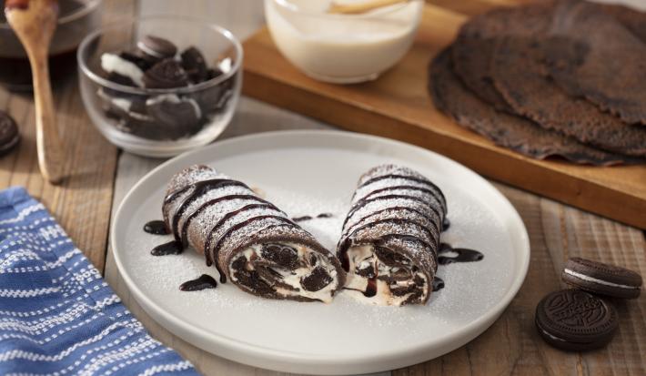 Foto da receita de Burrito Doce de Negresco, decorado com calda de chocolate e açúcar, em um prato branco grande, sobre uma bancada de madeira decorada com um tecido azul, alguns Biscoitos Negresco e dois poptinhos com caldas de chocolate
