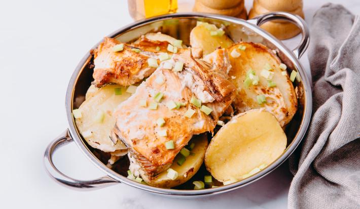 Fotografia de postas de peixe assado com molho à base de vinho e batatas. Por cima, cebolinha salpicada, o assado está dentro de um recipiente de alumínio fundo, que está sobre uma mesa de mármore ao lado de uma toalha na cor cinza.