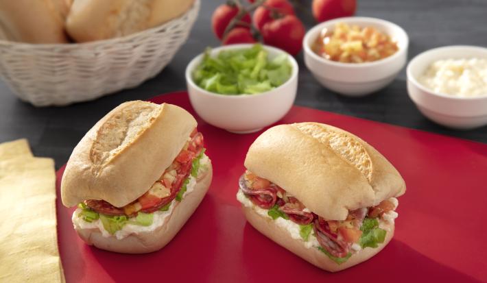 Foto de um sanduíche pequeno, recheado com uma creme branco, alface e tomate, sobre uma tábua vermelha em uma mesa com potinhos com ingredientes e um cesto de pães franceses ao fundo