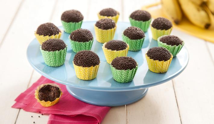 Fotografia em tons de marrom em uma bancada de madeira de cor branca. Ao centro, uma boleira azul contendo os brigadeiros. Ao lado, há um pano rosa com um brigadeiro em cima. Ao fundo, há algumas bananas.