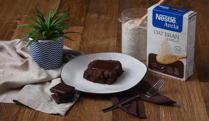 Fotografia de um pedaço de brownie com um quadrado de chocolate ao centro, sobre um prato branco com borda preta. O brownie está ao lado de uma embalagem de aveia farelo, sobre uma mesa de madeira.