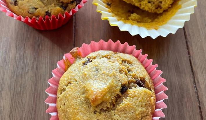 Fotografia de três forminhas de cupcake com bolo de cenoura e uva passa, sendo duas de tom rosa e uma amarela. Os cupcakes estão sobre uma mesa de madeira.
