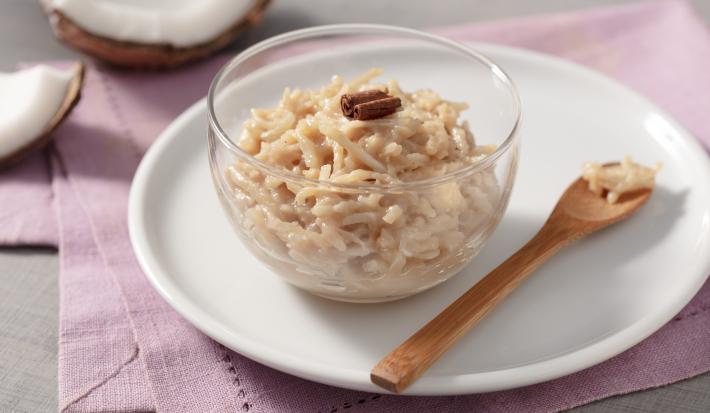Fotografia em tons de branco e rosa de uma bancada branca com um paninho rosa, sobre ela um prato redondo branco com um recipiente de vidro com cocada e uma colher. Ao fundo um coco.