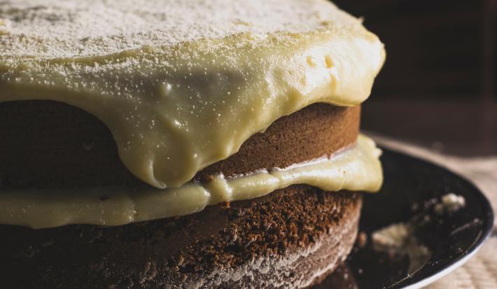 Fotografia de um bolo de abóbora com creme de coco por cima. O bolo está recheado com o mesmo creme, e está sobre um prato raso preto.