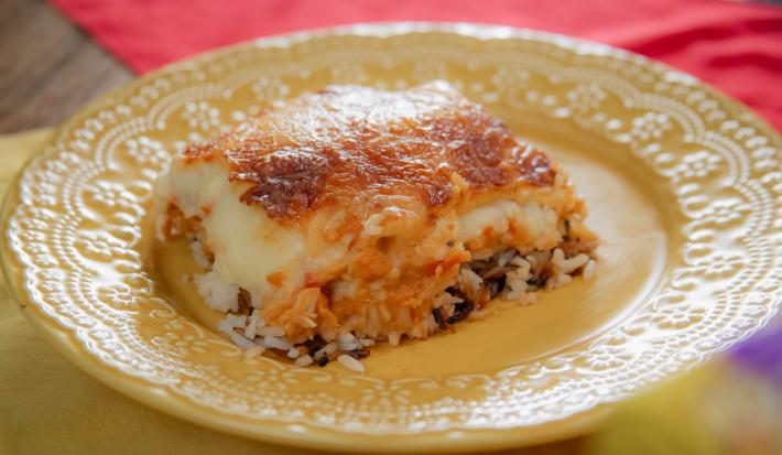 A fotografia mostra Escondidinho da Dona Rosa servido em um prato branco, exibindo camadas perfeitas de arroz soltinho, frango cremoso e purê de batatas dourado, com uma crosta de queijo parmesão e muçarela gratinados