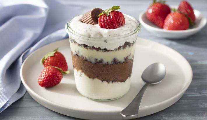 Imagem de um copo com a receita mostrando as camadas de creme, decorado com morango e um bombom. Ao fundo, um potinho com morangos.