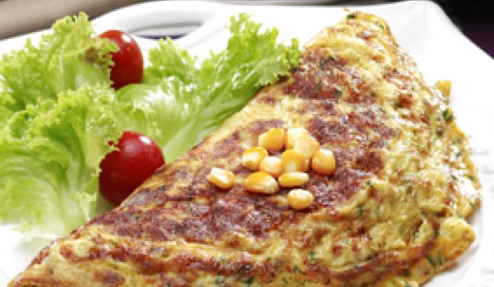 Fotografia em tons de verde em uma mesa com uma toalha listrada com marrom e bege, um prato branco redondo com uma omelete com milho e salada de alface e tomate cereja ao lado do prato.