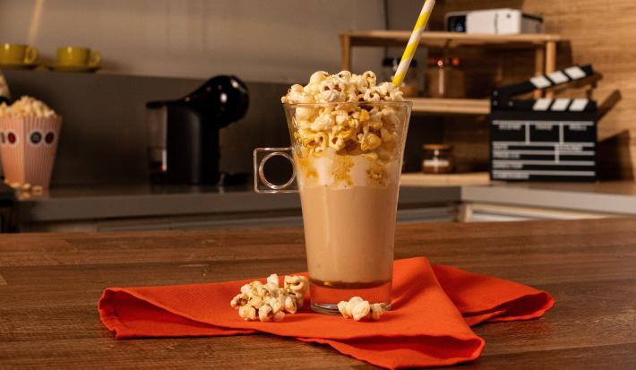 Fotografia de uma xícara de vidro com uma bebida de NESCAFÉ DOLCE GUSTO Netflix Caramelo Salgado com pipoca por cima e um canudo de cor amarelo e branco. O copo está apoiado sobre um pano vermelho, e está sobre uma mesa de madeira.