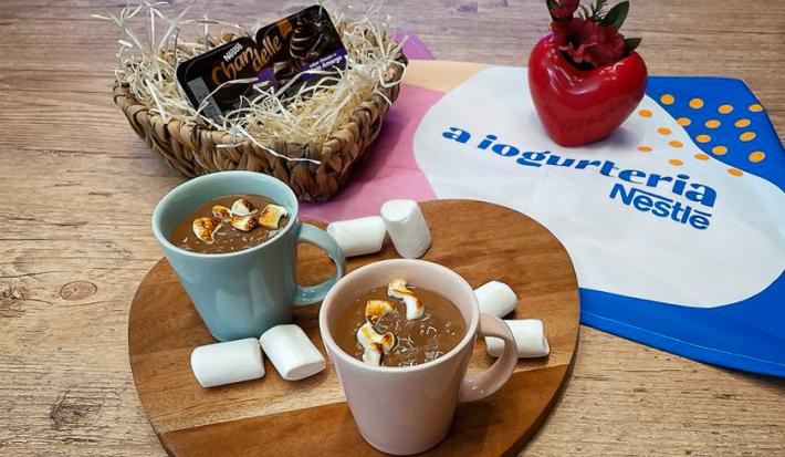 Fotografia em tons de marrom, em uma bancada de madeira de cor marrom. Ao centro, uma tábua de madeira contendo 2 xícaras com as bebidas. Ao fundo, há um pano de A Iogurteria Nestlé e um pote de chandelle ao lado.