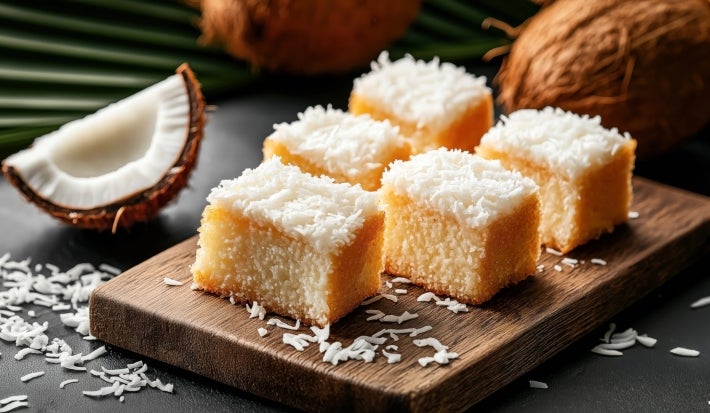 Fotografia de pedaços fofinhos de bolo de coco dispostos em uma tábua de madeira, com cobertura de coco ralado e uma crosta dourada.