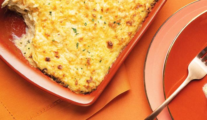 Fotografia em tons dourado e ferrugem, com refratário de cor ferrugem ao centro com escondidinho de bacalhau gratinado salpicado de cheiro-verde sobre guardanapo ferrugem, ao lado prato ferrugem com garfo e porção do escondidinho, sobre bancada ferrugem.