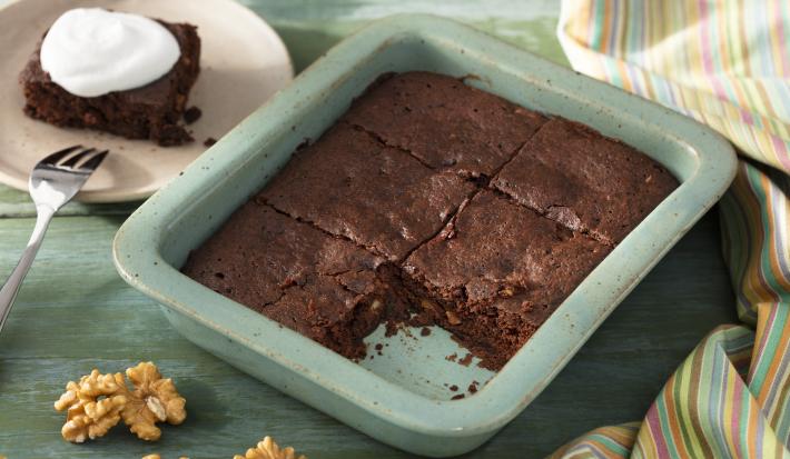 Fotografia em tons de verde em uma bancada de madeira verde, um recipiente verde quadrado com o brownie de chocolate com nozes e um pano colorido ao lado. Ao fundo, um prato redondo bege com uma fatia do brownie.