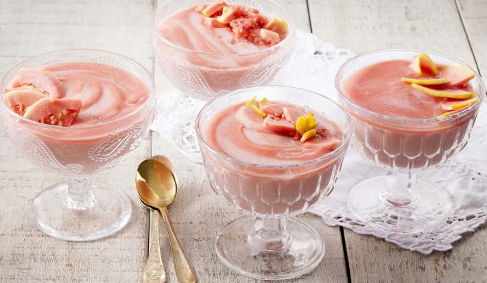 Fotografia em tons de rosa e branco, com 4 taças contendo mousse de goiaba decorada com fatias da própria fruta, sendo duas delas sobre guardanapo branco, duas colheres douradas ao lado, tudo sobre bancada de madeira com pátina.