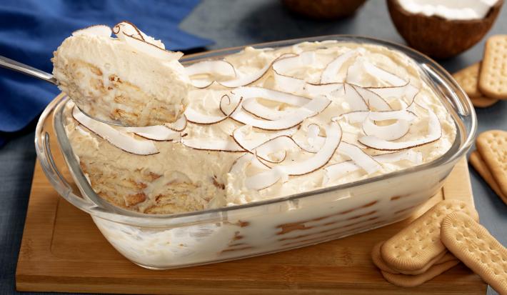 Fotografia em tons de branco, com tábua de madeira com um recipiente de vidro alto com o pavê de coco com biscoitos maisena São Luiz espalhados pela mesa.