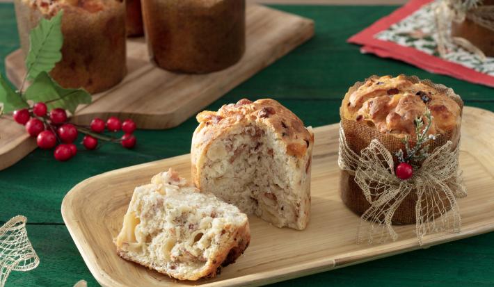 Fotografia em tons e bege em uma bancada de madeira de cor verde. Ao centro, uma bandeja de madeira contendo os mini panettones. Ao fundo, há mais alguns mini panettones em uma tábua de madeira.