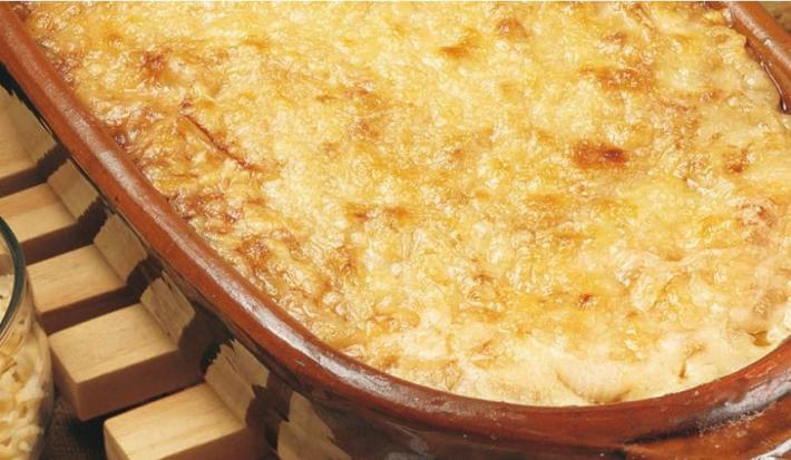 Fotografia em tons de marrom em uma bancada com um suporte para panela de madeira, uma travessa de barro com as cebolas gratinadas dentro dela e em cima do suporte. Ao lado, potinho com queijo ralado.