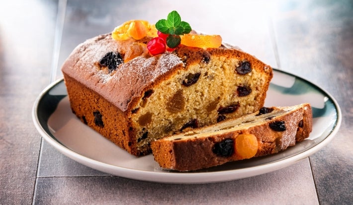 Fotografia de uma fatia de bolo de frutas em um prato, com uma crosta dourada e interior úmido, repleto de frutas secas. O bolo é decorado com açúcar de confeiteiro, folhas de hortelã e frutas cristalizadas.