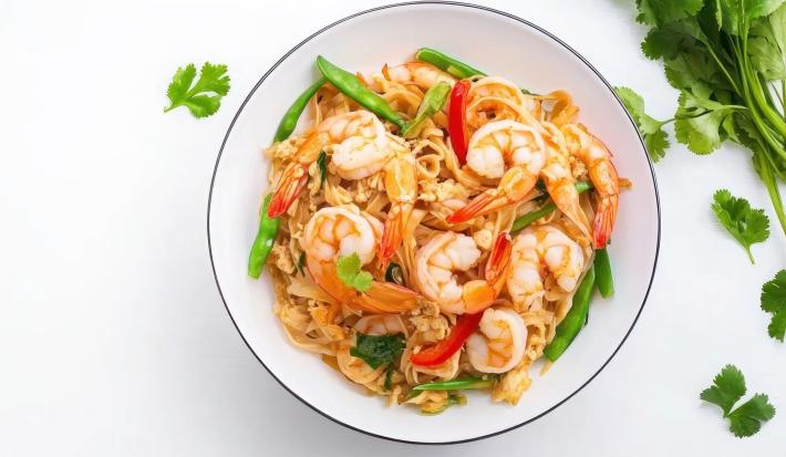 Fotografia vista de cima em tons claros de uma mesa branca. Sobre a mesa tem um prato fundo branco ao lado de um maço de coentro. Dentro do prato tem uma receita de pad thai com camarões.