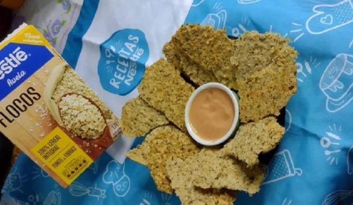 Foto da receita de crostini de aveia servida em diversas porções com um pote de molho ao centro. Isso sobre uma toalhinha de receitas nestlé e com uma embalagem de aveia flocos ao fundo