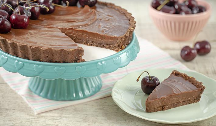 Fotografia em tons de marrom, branco, azul e rosa de uma bancada cinza vista de frente. Um recipiente azul contém uma torta de chocolate com varias cerejas e com uma fatia retirada, ao lado um prato branco com a fatia de torta e uma cereja do lado.