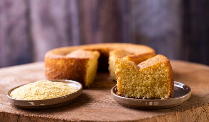 Foto da receita de bolo de milho de liquidificador, redondo, dourado, com uma fatia mais à frente em um prato e ao lado um punhado de fubá em outro prato. Ao fundo é possível ver o bolo inteiro