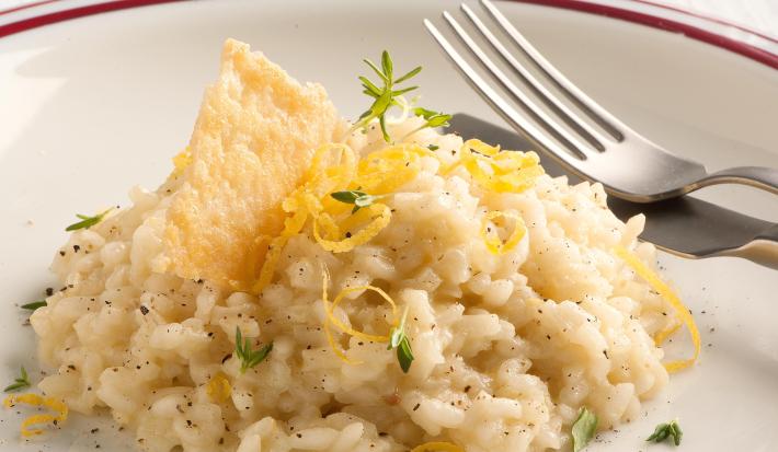Fotografia de um prato branco com uma listra vermelha na borda, onde está servido o risoto decorando com raspas de limão siciliano e uma crosta crocante de queijo parmesão. Ao fundo uma taça de vidro transparente e um guardanapo de tecido branco.