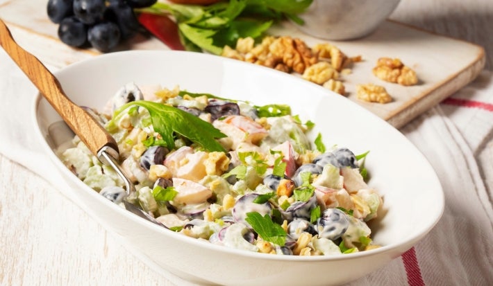 Fotografia de uma salada em uma tigela oval branca, com maçãs, melão e uvas pretas em um molho cremoso, decorada com nozes e salsa, acompanhada de um garfo, em um ambiente rústico com ingredientes frescos ao fundo.
