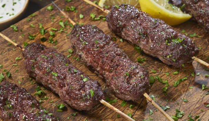 Fotografia de quatro espetinho de carne moída sobre uma tábua de madeira, e por cima, salsinha salpicada. Ao fundo, um molho de pepino dentro de um recipiente de fundo e pequeno, e ao lado, dois pedaços de limão siciliano.