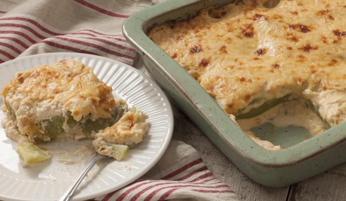 Fotografia em tons de branco e dourado, forma com Chuchu Gratinado sobre bancada de madeira rústica e prato branco com um garfo e uma porção do Chuchu Gratinado sobre guardanapo branco com listras de cor vinho.