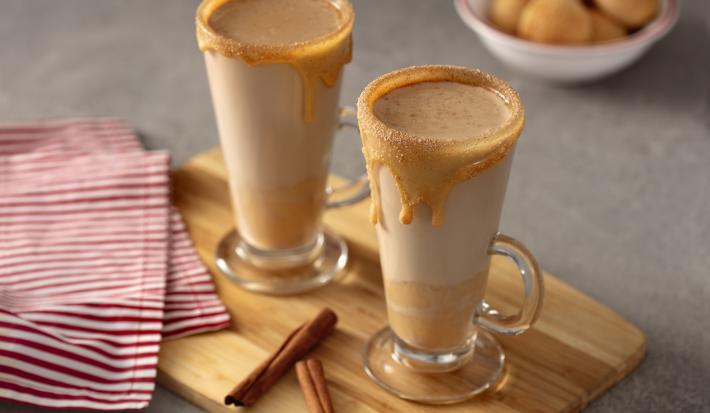 Foto de uma bancada. Sobre ela há uma tábua marrom com uma guardanapo de tecido listrado vermelho e branco, duas taças com as bebidas marrons dentro e com as bordas decorada com creme de avelã. Ao fundo há um pequeno potinho branco com pães de queijo