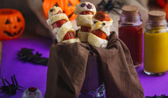 Imagem aproximada da receita de salsichas envoltas em tiras de massa folhada com pontinhos pretos, simbolizando olhos. Na cena há abóboras falsas, dois potes com ketchup e mostarda, algumas aranhas falsas, tudo numa superfície em tom roxo escuro
