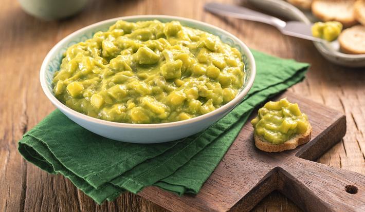 Foto da receita de Guacamole. Observa-se um pote redondo com a guacamole, sobre um guardanapo verde e uma tábua de madeira. Ao lado do recipiente, uma torrada redonda com o guacamole. As mesmas torradas decoram o fundo da foto.