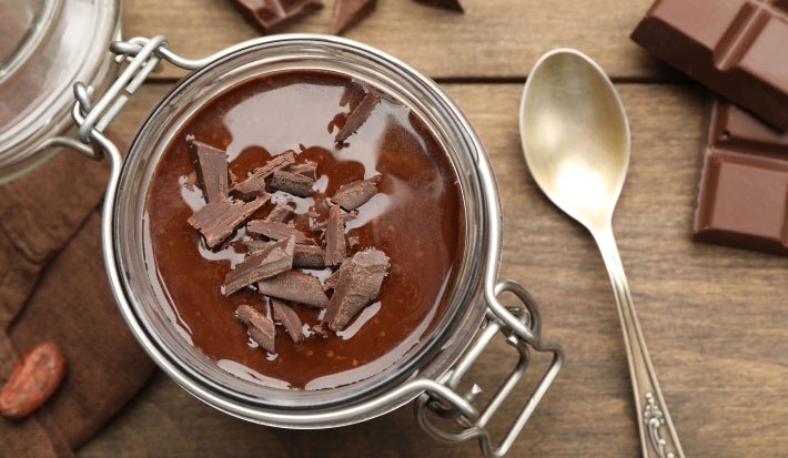 Fotografia vista de cima de um recipiente de vidro, tipo um copo, que fecha sobre uma mesa de madeira. Dentro tem a sobremesa, finalizada com ganache e raspas de chocolate, e ao redor tem uma colher pequena e alguns pedaços de chocolate.