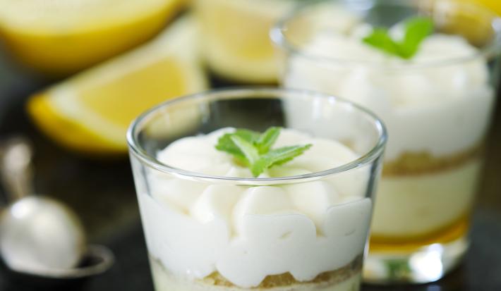 Fotografia de dois copinhos de vidro com farofa de biscoito e creme de limão sobre uma mesa de mármore preta. Ao fundo, duas metades de um limão siciliano, muitas folhas de hortelã e uma colher de sobremesa.