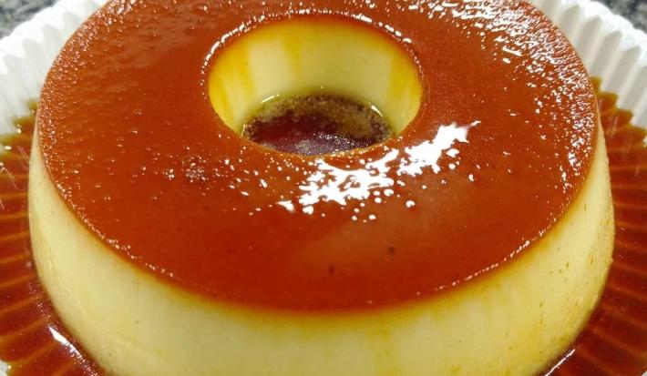 Fotografia em tons de bege em uma mesa de mármore, um prato branco redondo com o pudim de leite gourmet com calda de caramelo em cima dele.