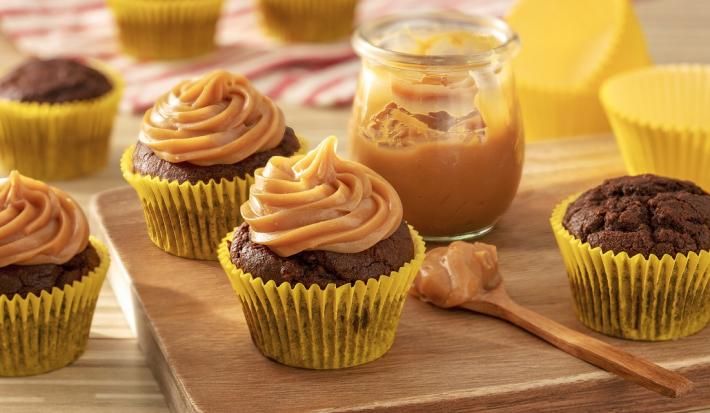 Fotografia em tons de amarelo em uma bancada de madeira de cor marrom. Ao centro, uma tábua de madeira contendo os cupcakes e um pote contendo o doce e leite com uma colher ao lado. Ao fundo, há um pano vermelho listrado.