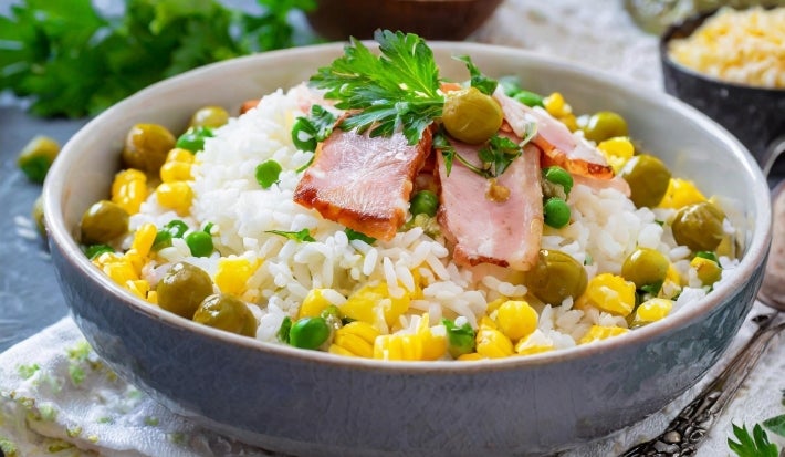 Em cima de uma mesa, tigela com porção de arroz branco soltinho com milho, azeitonas verdes, decorado com fatias de bacon e folha de salsa.