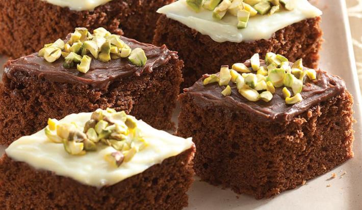 Fotografia em tons de marrom em uma mesa de madeira com uma toalha de renda, um recipiente branco retangular com os bolos de chocolate com cobertura e pistache.