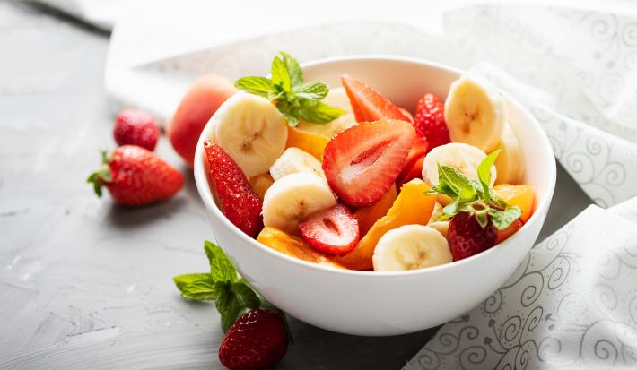 Fotografia de um recipiente pequeno e branco de frutas sobre um purê de banana e folhas de hortelã. O recipiente está sobre uma bancada de cor escura, do lado das frutas tem alguns morangos e mais folhas de hortelã.