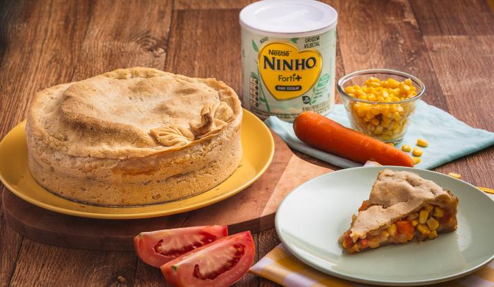 Fotografia em tons de amarelo e verde de uma bancada de madeira, sobre ela um prato amarelo com uma torta, a frente um prato verde com uma fatia da torta, mostrando o recheio de milho, cenoura e tomate. Ao fundo uma lata de Ninho Origem Vegetal.